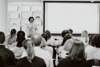 A black and white photo features Founder Chisom speaking in a classroom to a group of individuals.